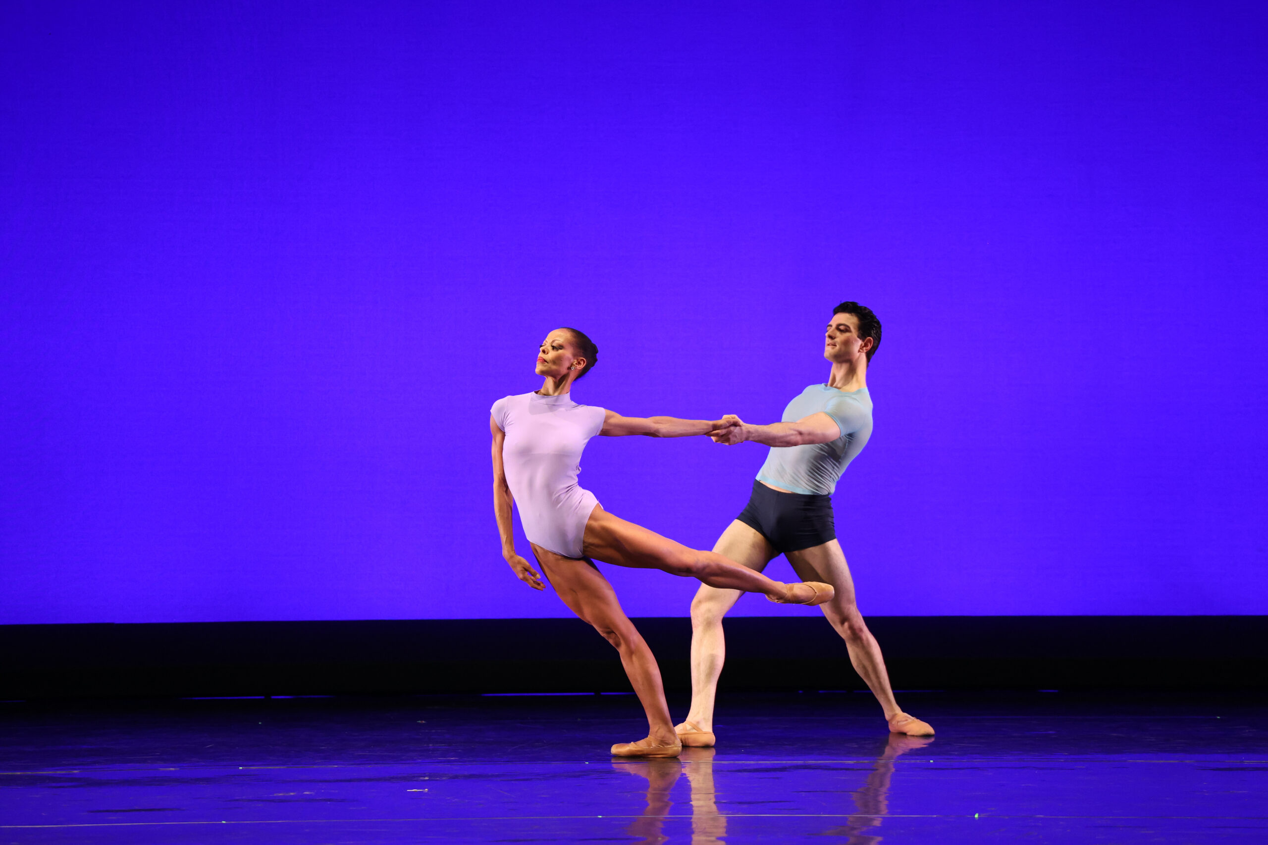 Myrna Kamara, wearing a lavender-colored leotard and brown pointe shoes, does a low arabesque, leaning into her right hip as her partner, Ariel Merkuri, pulls her left hand behind her. Merkuri wears a light blue top and dark shorts, and they perform in front of a bright blue backdrop.