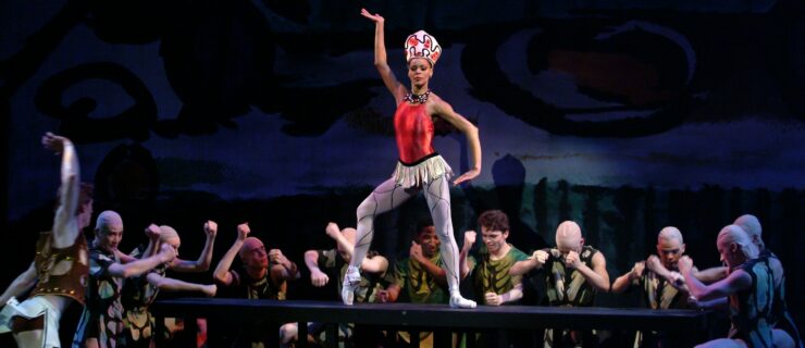 Myrna Kamara, costumed in a red bodice, shirt white skirt, white tights and pointe shoes, and a large white and red headpiece, stands on a table and props her right foot up onto pointe. She raises her right arm high, flexing her hand out, and her left arm low, with the hand flexed in. A large corps of male dancers in green and brown costumes surround her on three sides and pound the table wit their fists.