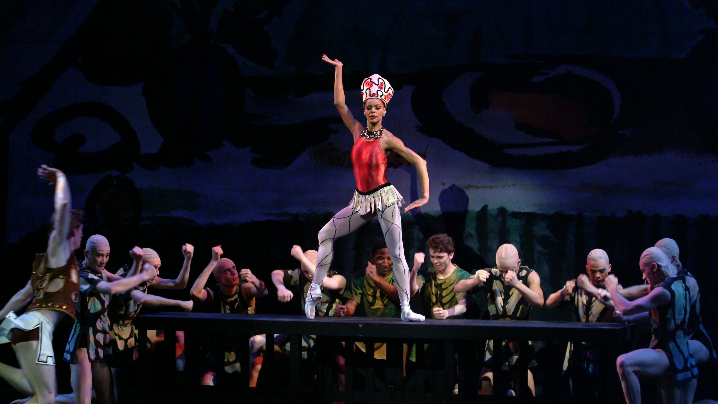 Myrna Kamara, costumed in a red bodice, shirt white skirt, white tights and pointe shoes, and a large white and red headpiece, stands on a table and props her right foot up onto pointe. She raises her right arm high, flexing her hand out, and her left arm low, with the hand flexed in. A large corps of male dancers in green and brown costumes surround her on three sides and pound the table wit their fists.
