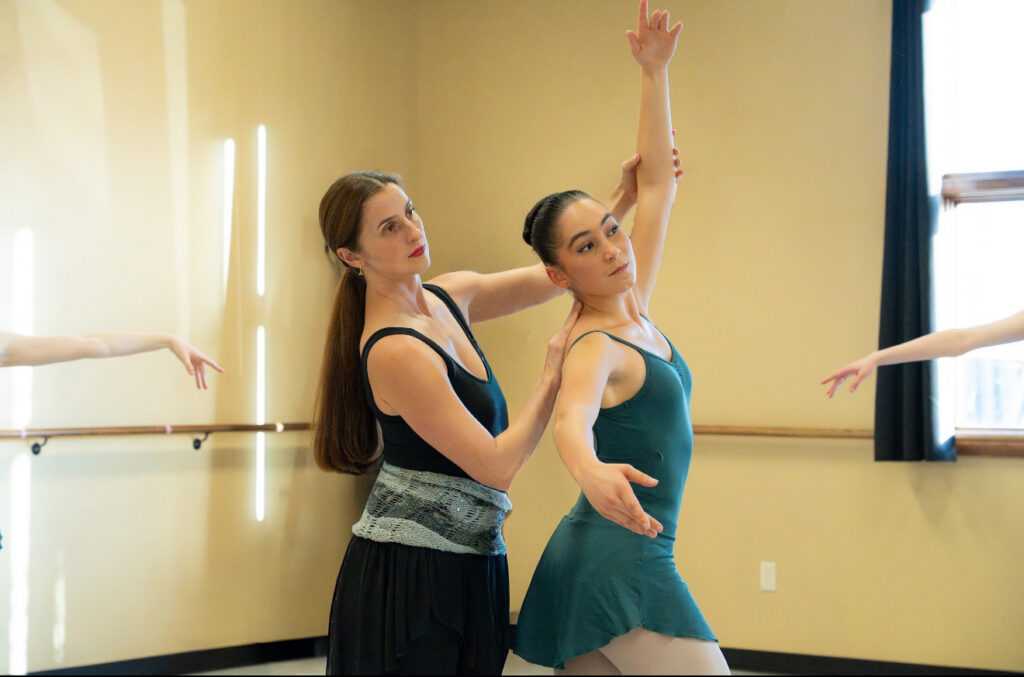 Alison DeBona, wearing rehearrsal clothes, adjusts the back and left arm of a teenage ballet student during a ballet class. The student wears a green leotard and short chiffon skirt, and pink tights.