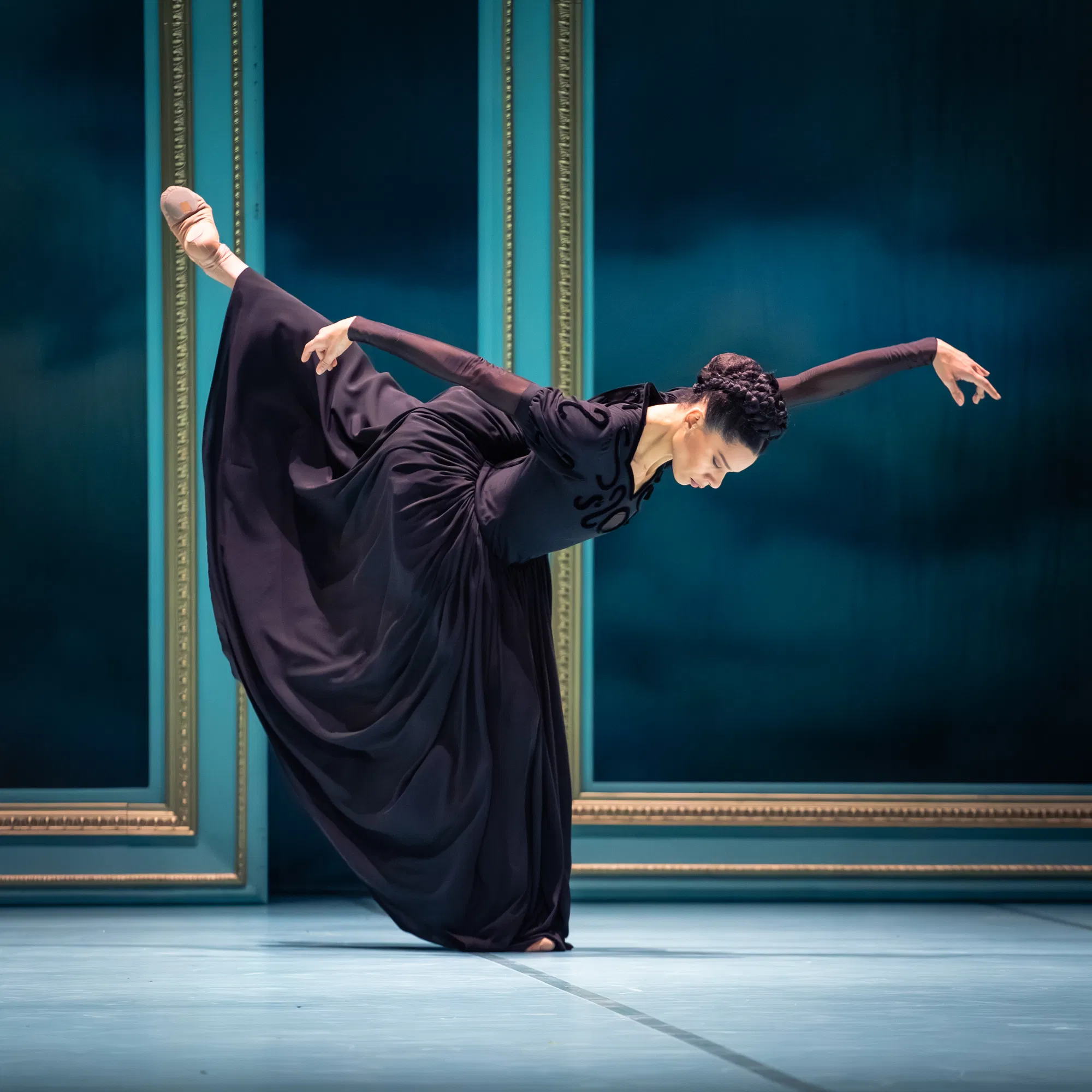 Onstage in front of a striking teal backdrop with gold accents, a dancer poses in arabesque with her supporting leg slightly bent. She bows forward with her arms in line with her upper body, in a long arabesque line. She wears a long, dark purple dress with a full skirt.