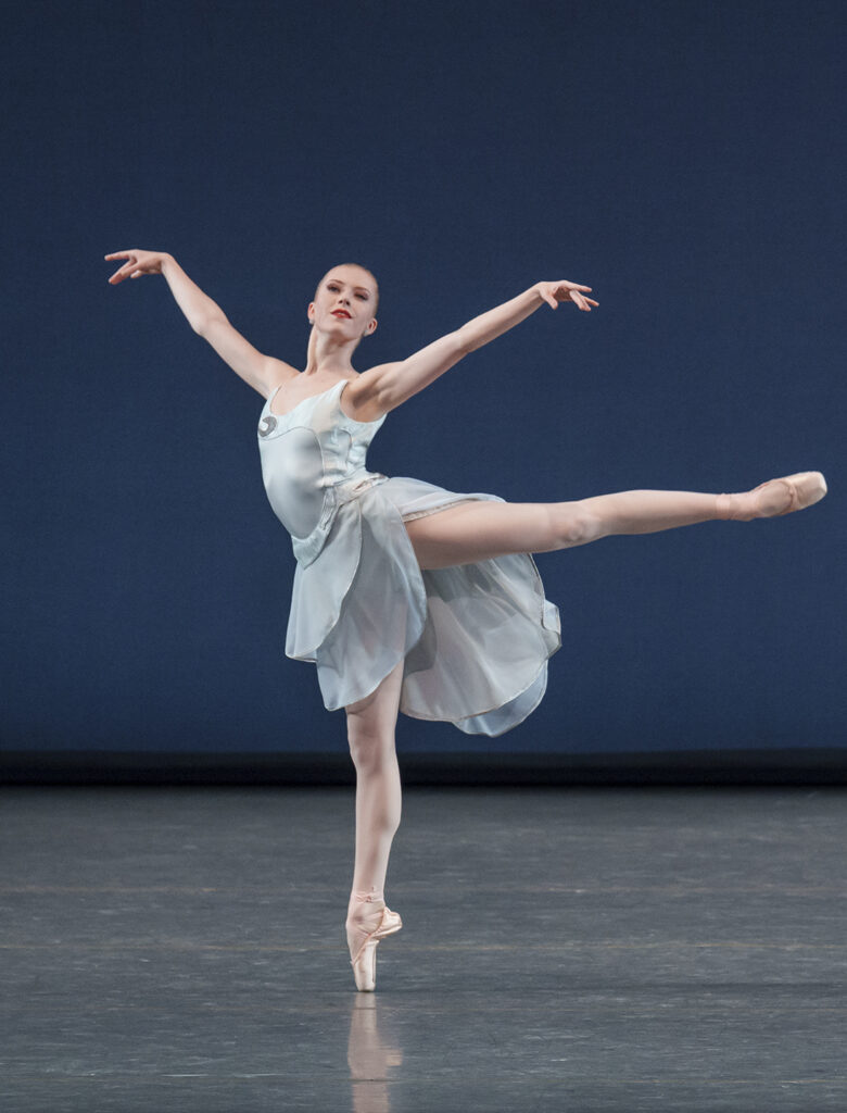 Onstage in front of a dark blue backdrop, a ballet dancer does an arabesque on pointe with her arms floating up to the side. She wears a light blue dress with a high-low effect and layers.