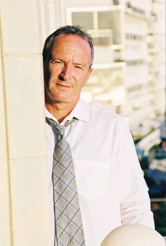 Michael Vernon's headshot. He stands partially hidden behind the edge of a building, wearing a white button-down and a gray scarf with blue lines.