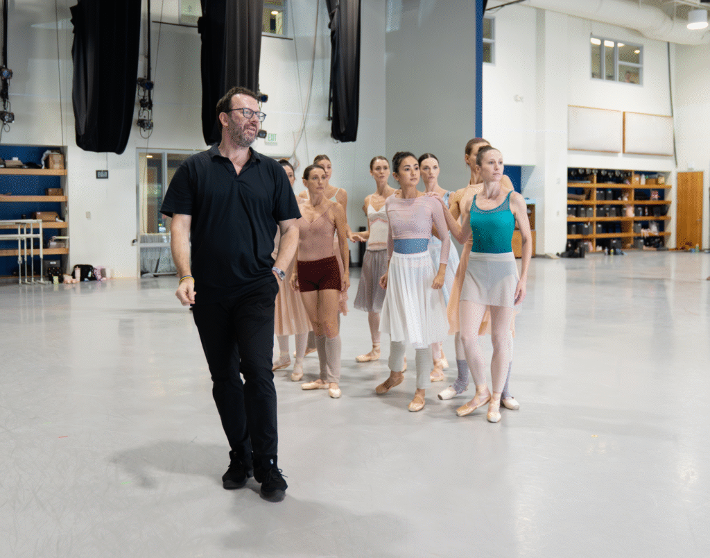 In a large ballet studio, a choreographer dressed in all black faces stands in front of a group of dancers, who are arranged in a clump. The dancers stand energetically, looking in the same direction as the choreographer, as if poised to move.