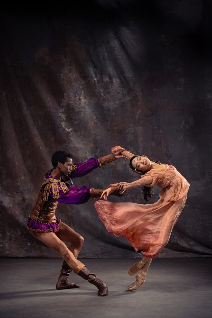 Calvin Royal III, is in profile facing his left; he pliés on his left leg and stretches his right leg forward for balance as he pulls his partner, Fangqi Li. Li poses in sur le coup de pied dérriére on pointe and arches back, looking out over her right should and smiling, as Royal III pulls her arms. She wears a billowing pink dress, while Royal II wears a purple and gold tunic, tan tights, and brown ballet boots.