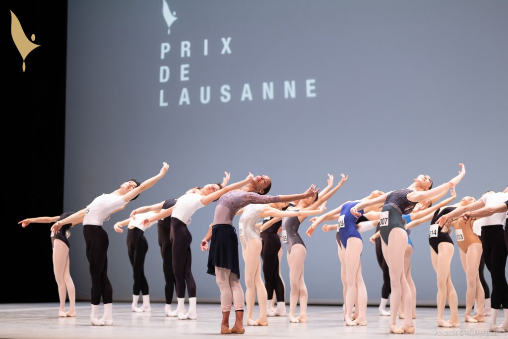 A ballet teacher leads an onstage class of teenage ballet students, They all stand in fifth position croisé and do a cambré back with their left arms up. The teacher wears a purple leotard, black skirt and pink tights rolled at the ankle, while the students wear practice clothing and have numbers pinned to their torsos.