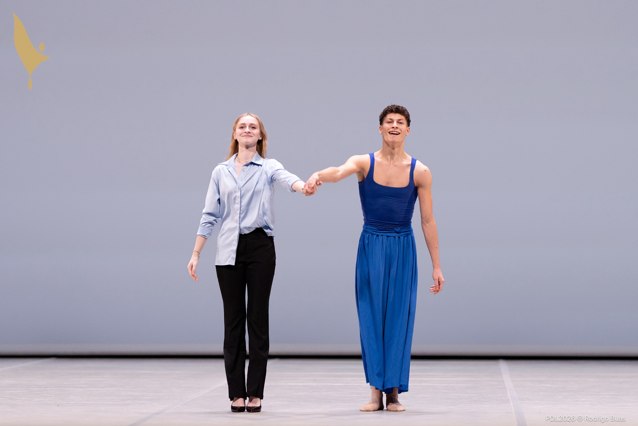 A teenage girl in a blue button-down shirt and black pants holds the hand to a male dancer to her left costumed in a blue tank top and wide-leg pants. They smile and prepare to take a bow onstage.
