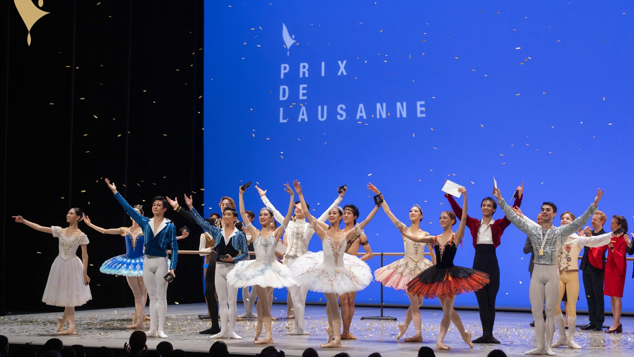 Fourteen teenage dancers in various costumes take a bow onstage as confetti rains down on them. Other dancers and adults stand towards the back of the stage, applauding. A blue backdrop says "Prix de Lausanne" in the upper corner.
