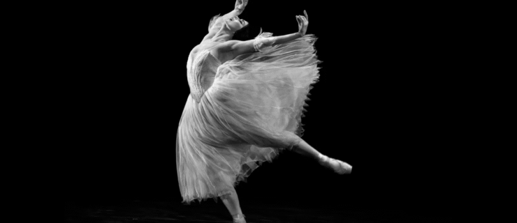 A black and white photo of Natalia Osipova, who dances with abandon onstage. She leans forward with one leg lifted in a low arabesque, her supporting leg slightly bent, as she arches and lifts her arms up and back. Her eyes closed, she opens her mouth with emotion. She wears a Romantic tutu with wispy arm puffs; the skirt of the tutu flies out behind her.