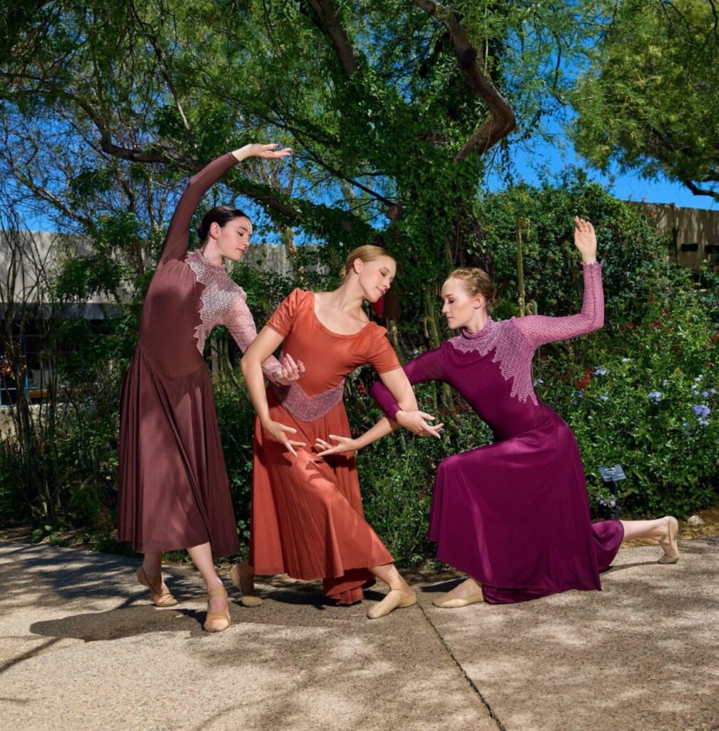 Outdoors, in front of a large tree, three dancers pose with interlocking arms. They wear long dresses in brick, orange, and magenta.