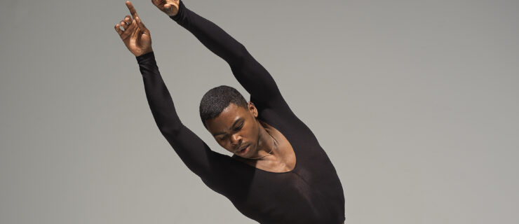 Against a gray ombre background, Sam Akins, a Black male dancer in a long sleeved black top, is shown from the waist up with his hands over his head and leaning towards the left.