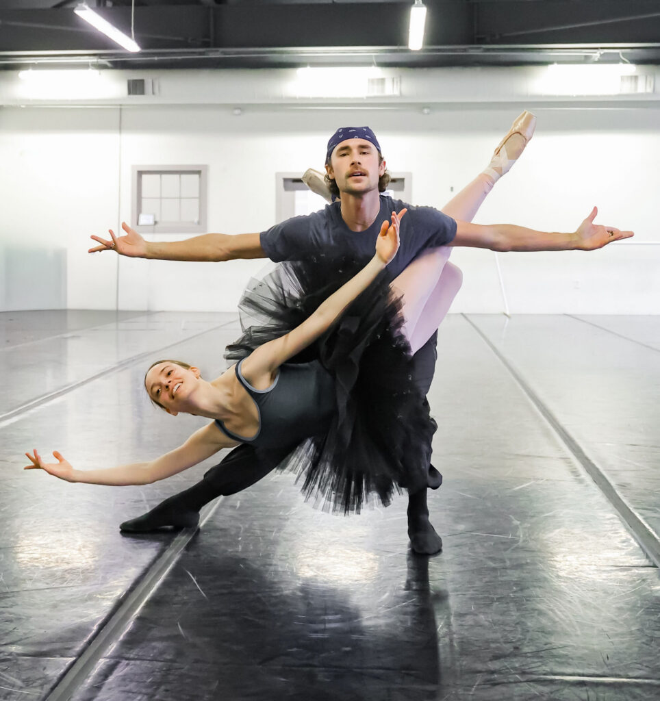 In a ballet studio, two dancers rehearse a "fish" in a pas de deux. They both extend their arms out triumphantly.
