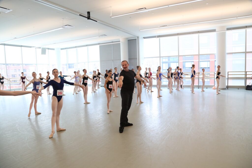A male ballet teacher stands at the front of a ballet class in a large studio and demonstrates a tendu à la seconde with his arms opening in second position. A large class of teenage dancers wearing leotards, pink tights, ballet slippers, and numbers pinned to their torsos stand behind him, learning the combination.