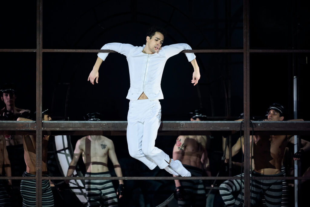 In a jail scene, David Soares, as Rudolf Nureyev, bends his arms to hang from a metal bar underneath his elbows. He bends his legs and turns them to the left slightly, looking down to the left with a pained expression. Behind him, a group of shirtless men in gray and black striped pants and policeman hats walk around and watch him.