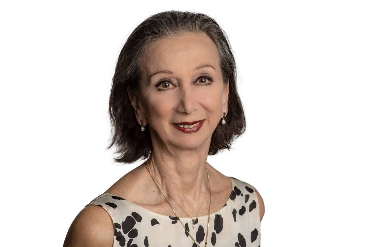 Maina Gielgud is shown from the chest up, She wears an off-white sleeveless top with black spots and two gold necklaces, and smiles directly at the camera. She poses in front of a white background.