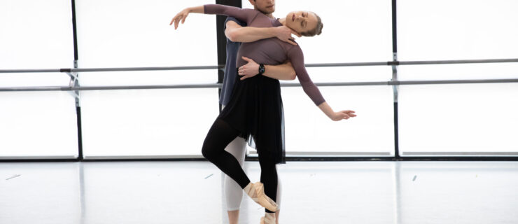 In a bright ballet studio, two dancers rehearse a sensual pas de deux. The dancer in front stands with one leg on pointe, the other in cou de pied devant, while leaning sideways into her partner's arm. She lets her head drop to the side, eyes closed and lips parted. Her partner stands behind her, wrapping one arm around her waist, with the other reaching across to grab hold of her neck.