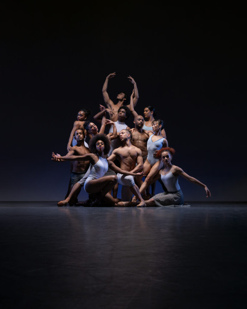Lit with a spotlight in front of a black backdrop, the company dancers of Ballet Black pose in a layered formation for a group photo. They all wear light blue or white dancewear.