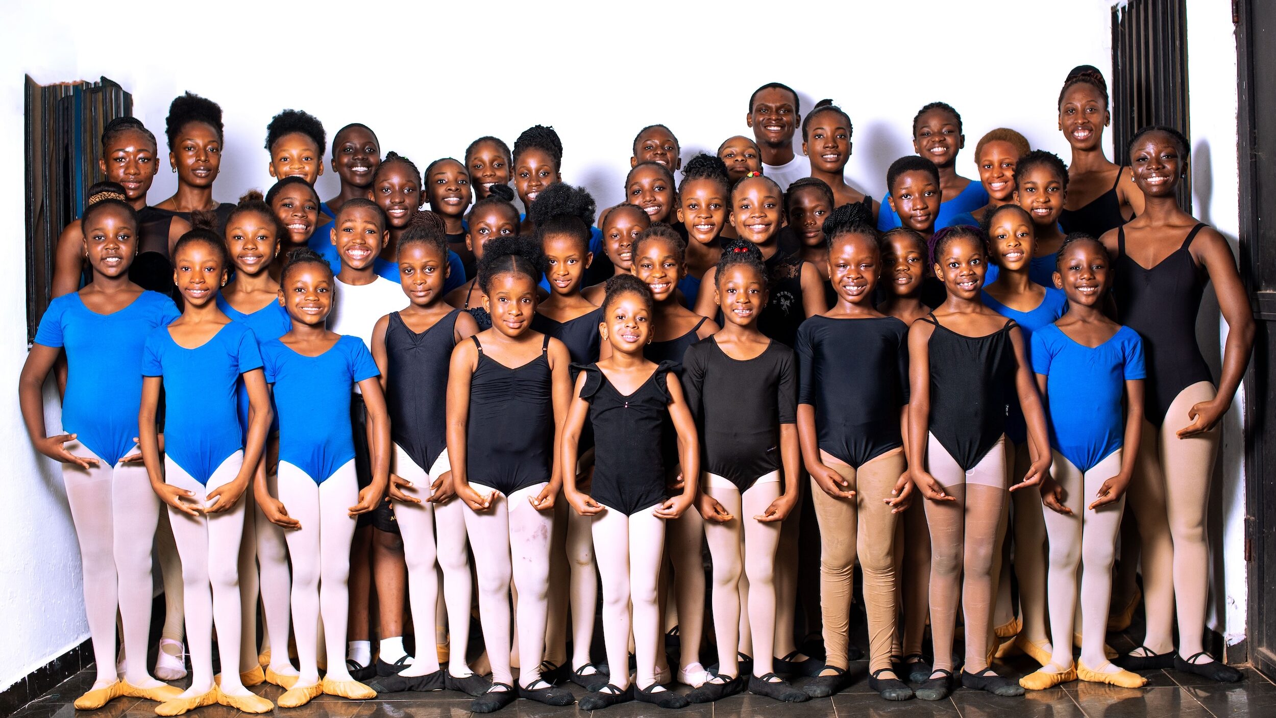 A large group of ballet students wearing leotards, tights, and ballet slippers stand in a large cluster, posing in first position with large smiles.
