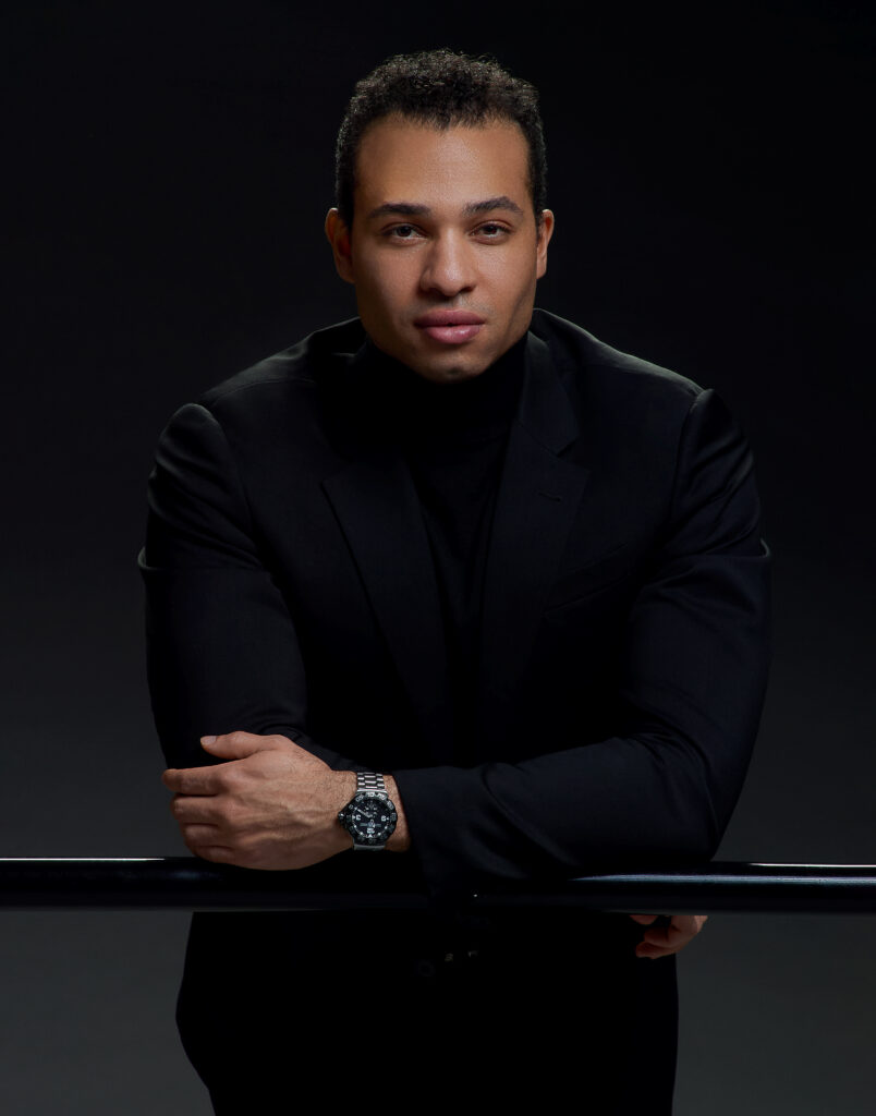 Adrian Blake Mitchell's headshot. He wears all black and leans forward while resting his forearms on a barre.