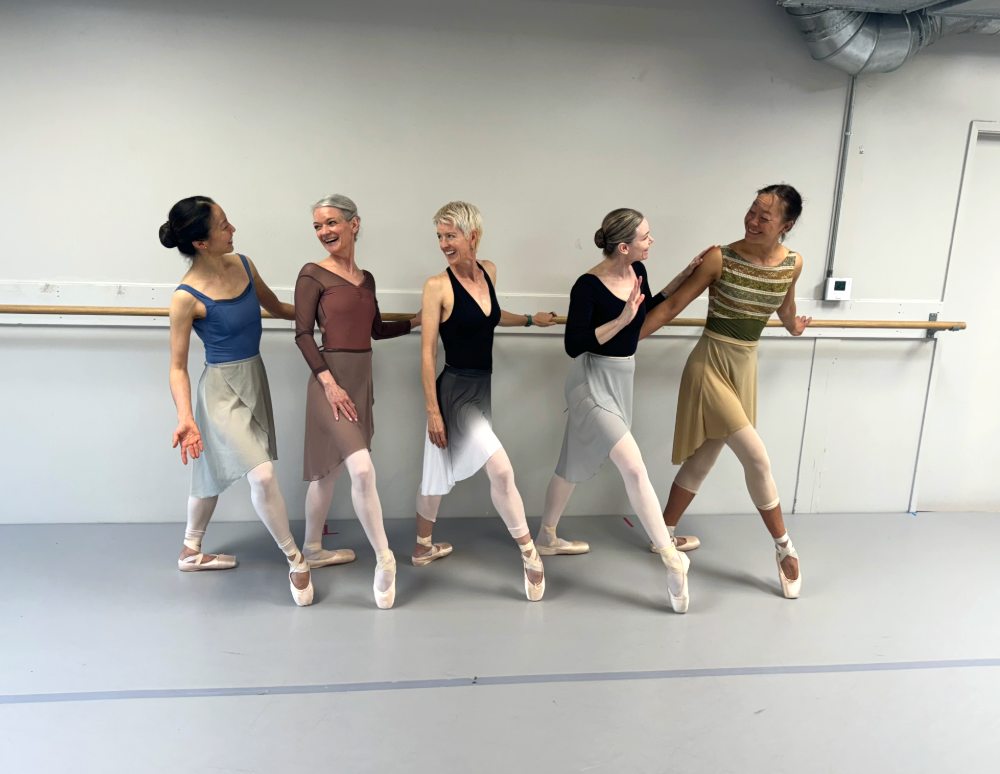 Five adult women wearing leotards, ballet skirts, tights, and pointe shoes pose along a ballet barre in a dance studio. They all bend their right leg, propping their foot up onto pointe, and hold onto the barre as they look at each other and smile.