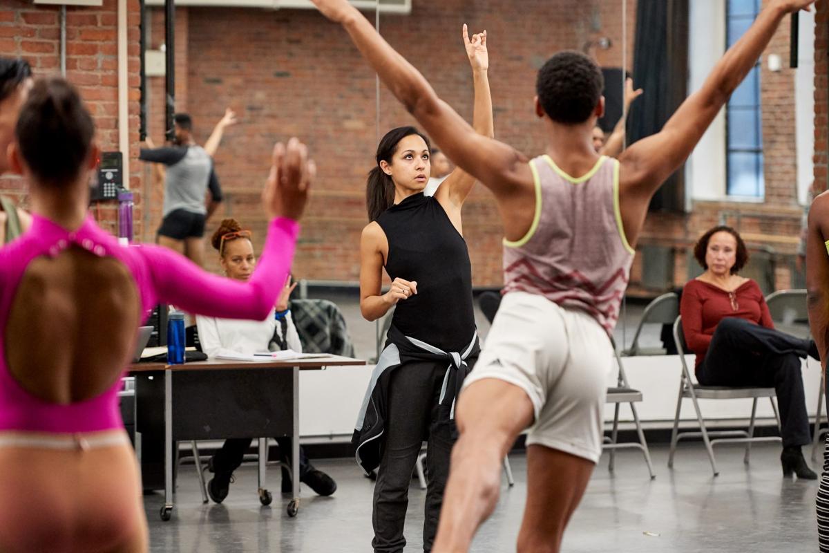 Claudia Schrieier, wearing a black leotard, black pants, and a sweatshirt tied around her waist, stands in the center of a studio as dancers practice ein front of her. She points upward with her left hand and looks up.