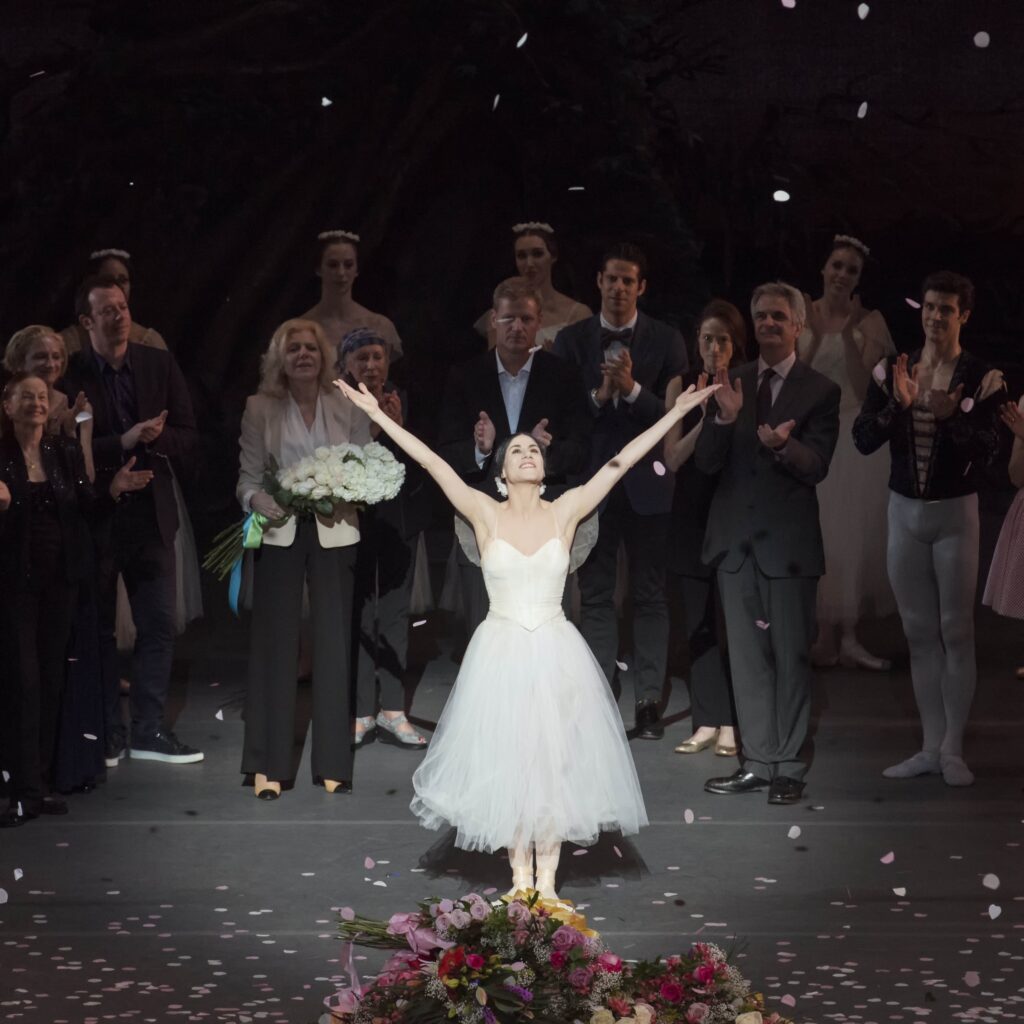 Paloma Herrera wears a white Romantic tutu and raises her arms in a final bow onstage. Bouquets of flowers sit at her feet and company members cheer behind her.