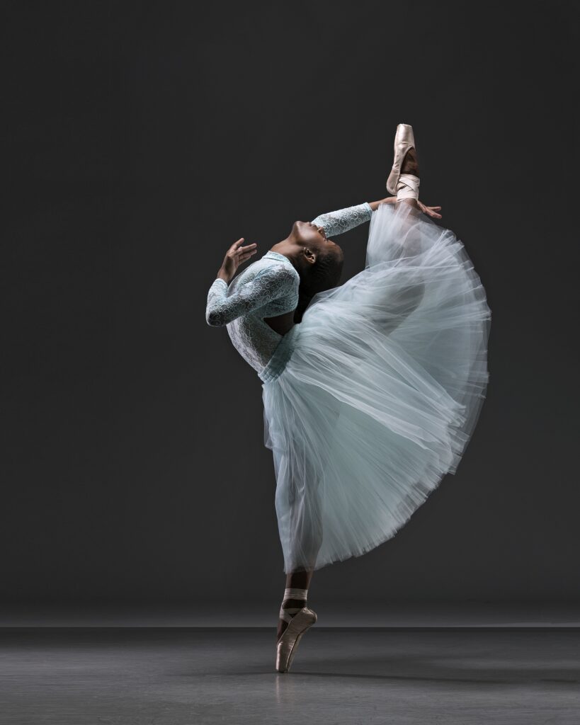 Marja’ Quaqua stands on pointe and kicks her leg in attitude dérriére. She arches back to meet her leg and wears a lacy blue leotard and Romantic tutu.