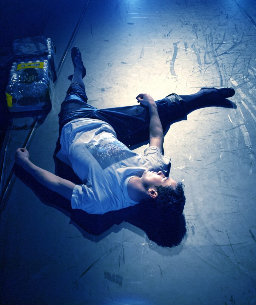 Cory Stearns lies flat on a marley floor next to a pack of water bottles. He wears a t-shirt, warm-up pants, and ballet slippers.
