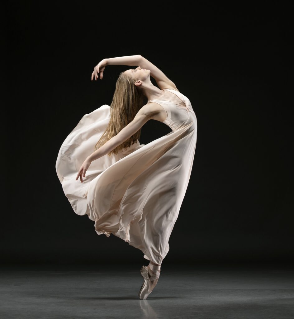 Annalisse Veldhuyzen VanZanten stands in profile in sixth position on pointe. She arches back, falling slightly forward, and wears a long, flowy light-pink dress.