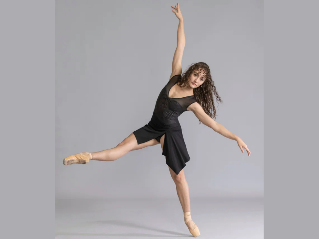 A dancer leans off-pointe with one leg lifted in a low devant position. Her arms extend on a high diagonal, and her curly hair swishes as she looks at the camera. She wears a black leotard and wrap skirt. 