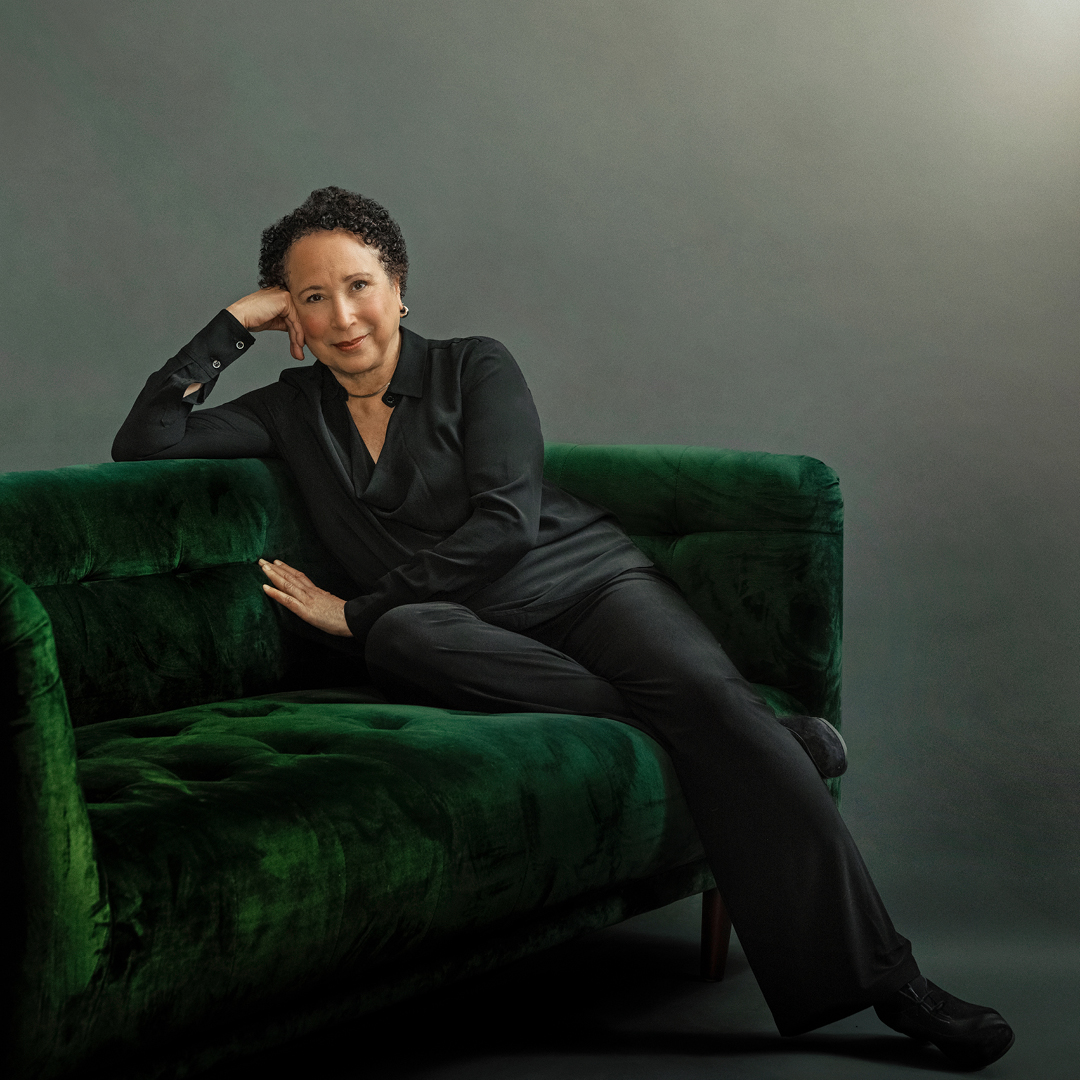 Virginia Johnson, wearing a dark suit, sits on a plush green velvet couch. She rests her head on her right hand as her arm extends along the top of the couch.