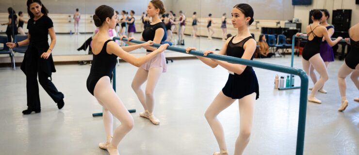 A trio of teen ballet students are shown facing a portable barre, with two on one side and one on the other, doing a demi plié in first position. They wear black leotards, short ballet skirts, pink tights, and pointe shoes. A teacher walks by and other dancers in practice clothes are shown in the background at various barres, doing the same thing.