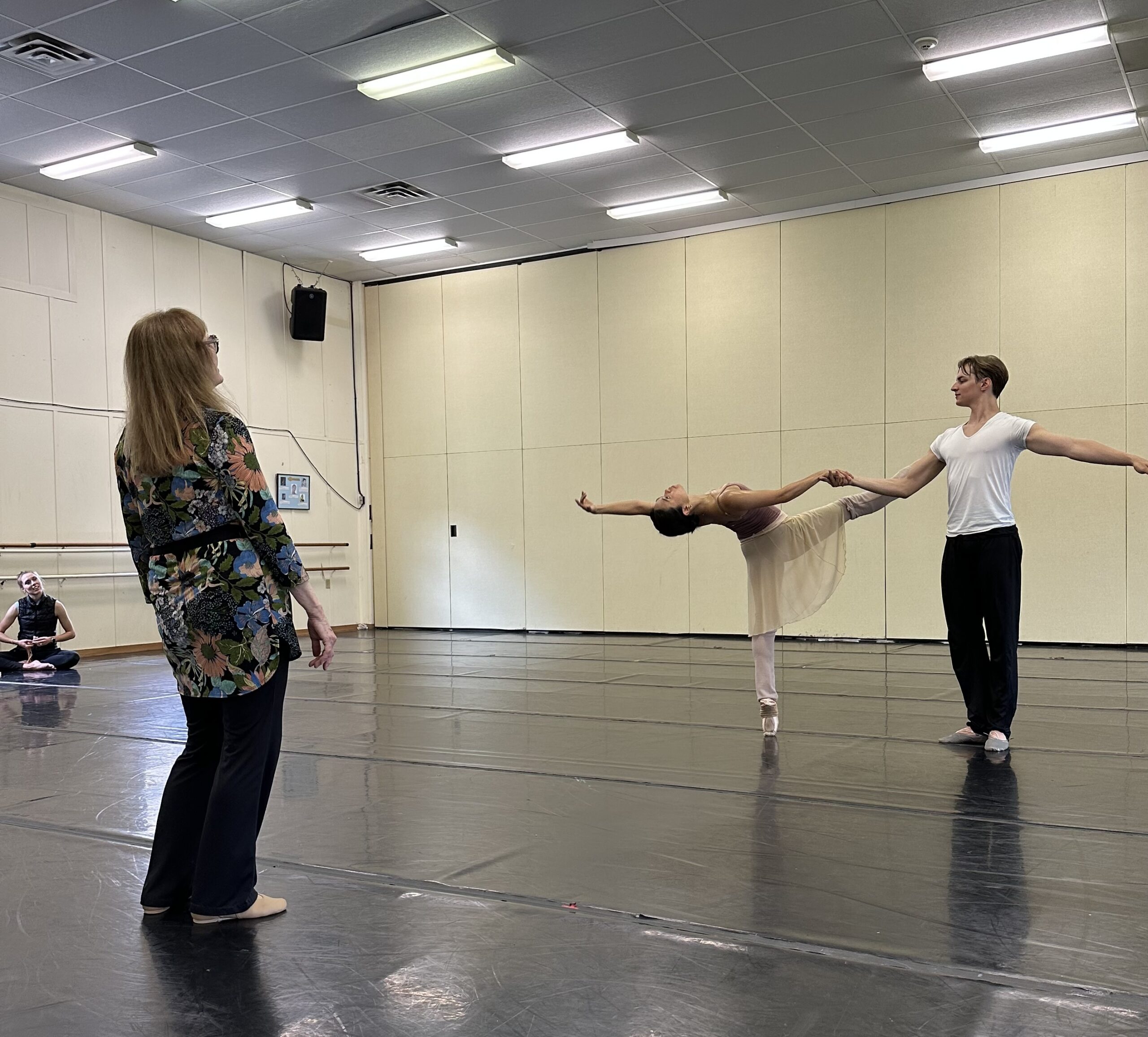 Patricia McBride is shown from behind as she rehearses two dancers in a pas de deux. She wears a filmy flowered tunic and black pants, while the dancers wear practice clothes.