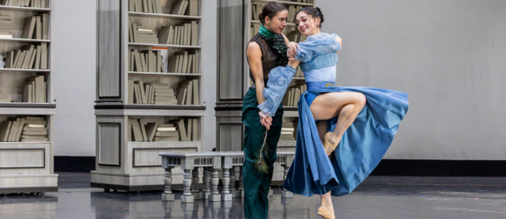 In a large ballet studio, two female dancers rehearse a pas de deux in costume. One wears dark green pants and a black shirt with a green ruffled collar. The other wears a blue dress with puffy sleeves and a slit down the side. Behind them are three large bookcase sets and some stools.