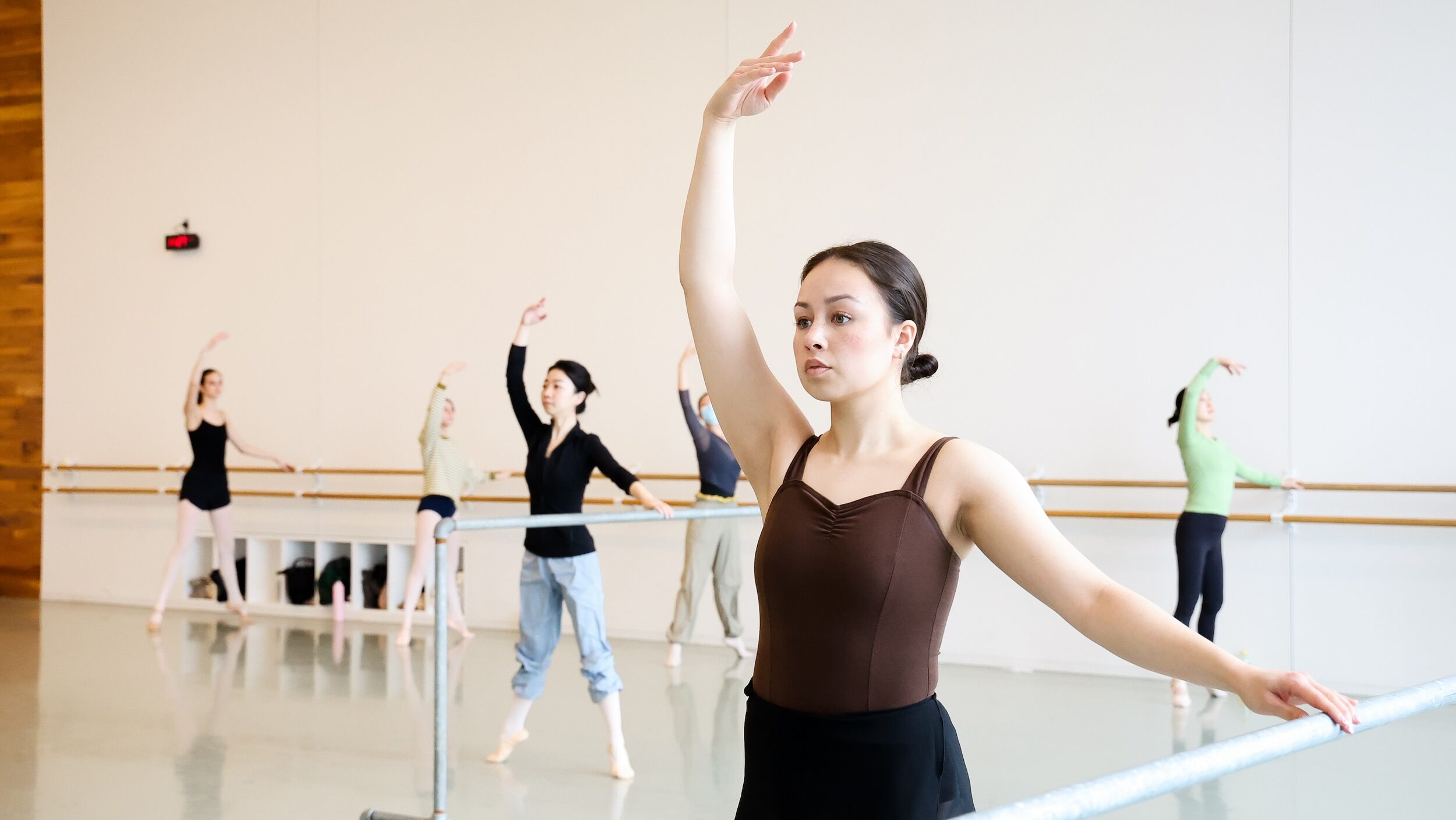 A dancer in a brown tank leotard and black skirt holds the barre with her left hand and lifts her right arm in fifth. Other dancers stand behind her at various barres, doing the same pose in écchappé second.