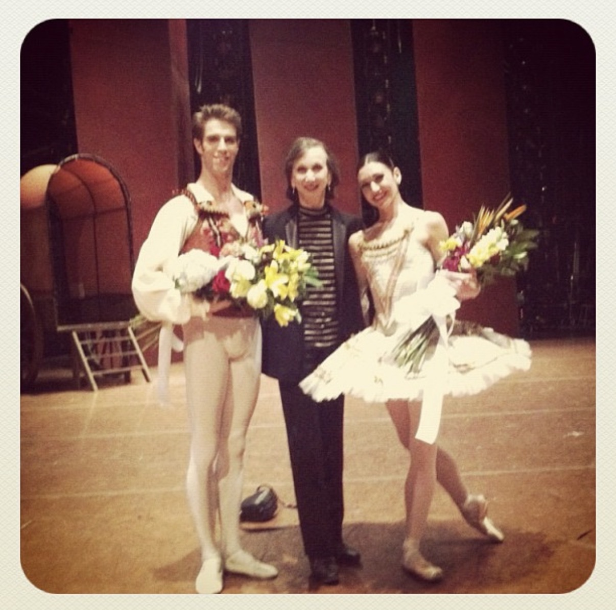 A male and female ballet dancer, wearing costumes, pose alongside their coach backstage. Both dancers hold bouquets of flowers.