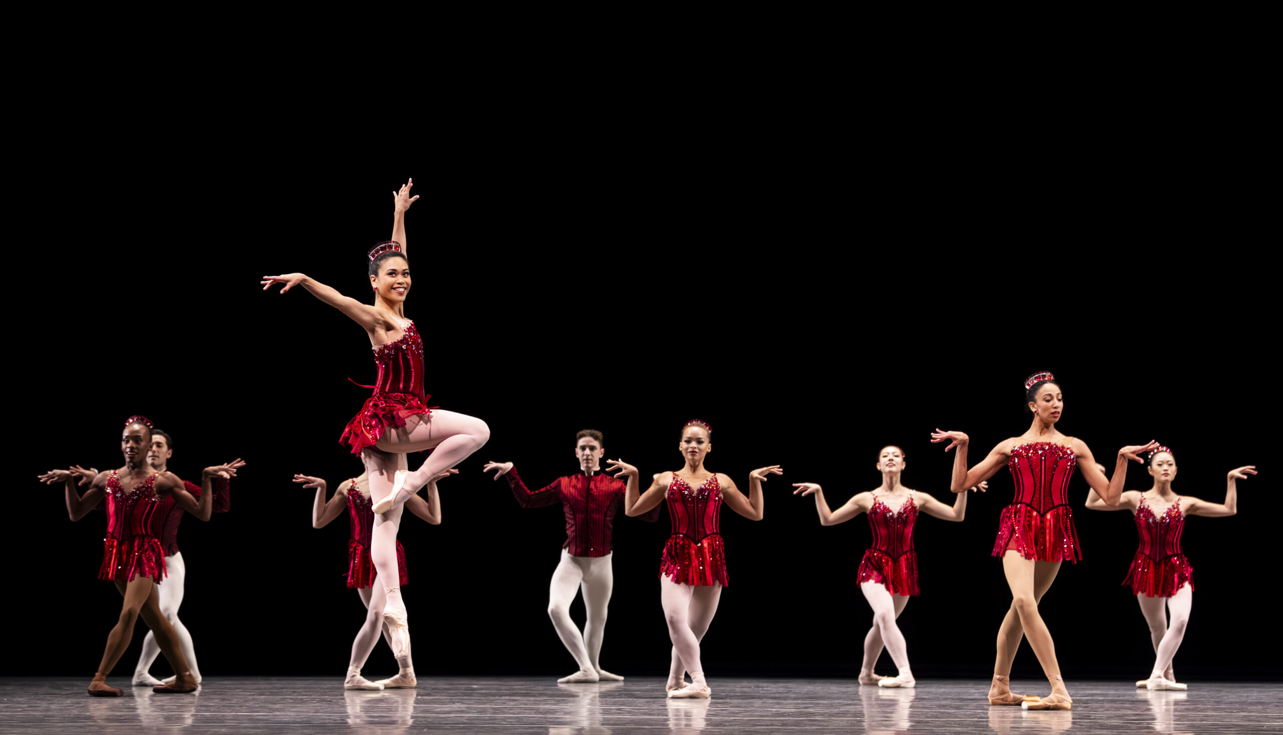 Angelica Generosa, wearing a red, bejeweled costume with a short red skirt, jumps up with her right leg in a parallel passé. A corps of dancers in red, bejeweled lunge in a staggered line behind her.