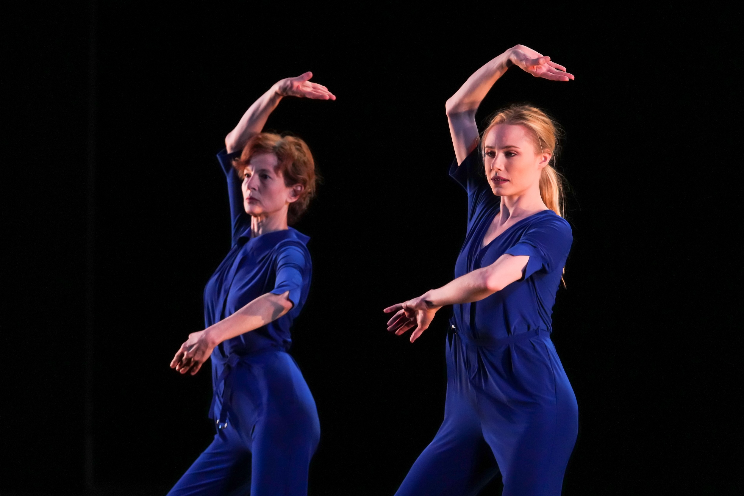 Two women costumed in royal blue pantsuits dance side by side.