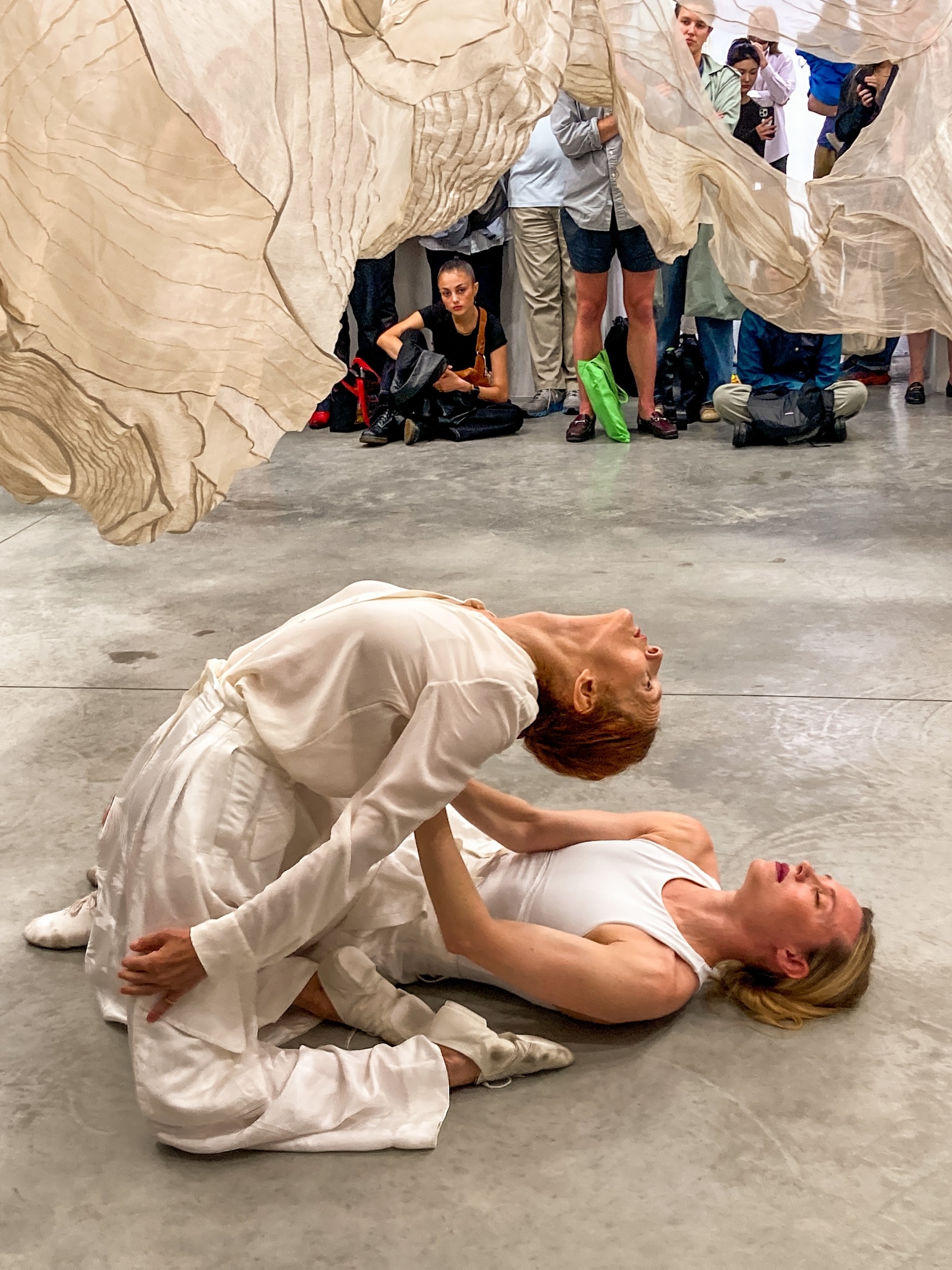 Sara Mearns lays on the ground as Jodi Melnick kneels on her left side and arches back. They both wear white costumes and dance under swaths of fabric as people sit and stand nearby, watching.