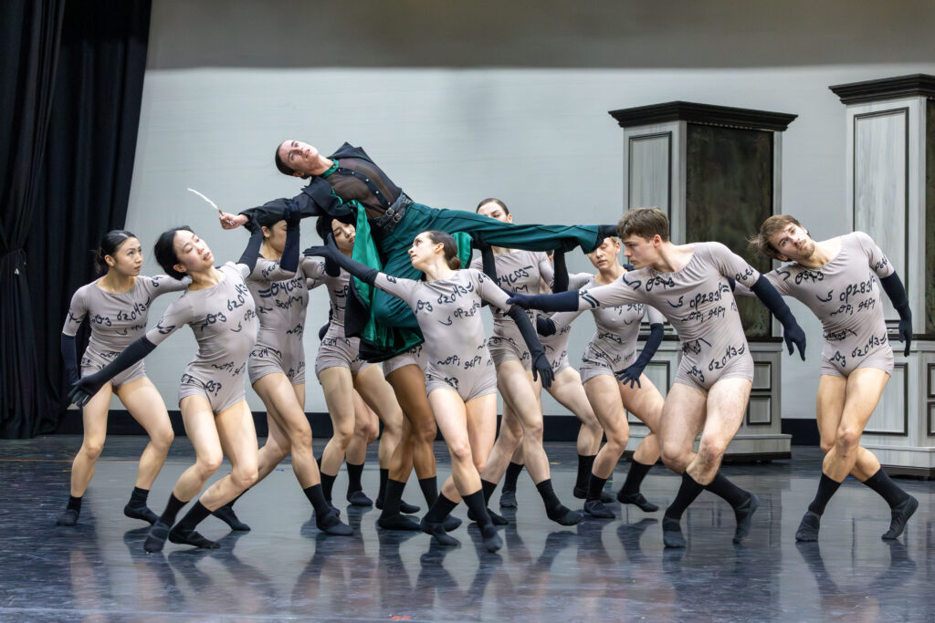 In a ballet studio, a large group of dancers in gray biketards lift another dancer wearing green slacks, a sheer black button-down, and a long black coat with green inner lining. She holds a quill and tucks one leg into retiré, her other extended to the side as she leans away from it. The dancers' biketards are printed with black calligraphy.
