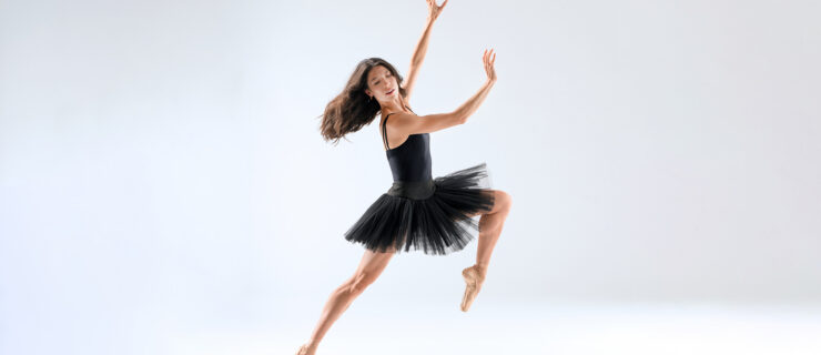 Charlotte Nash, wearing a black loetard and black tutu, leans off pointe on her right leg, her left leg bent and lifted. Her arms are held out in front of her, wrists flexed, as she looks down over her right shoulder, her long brown hair flowing behind her.