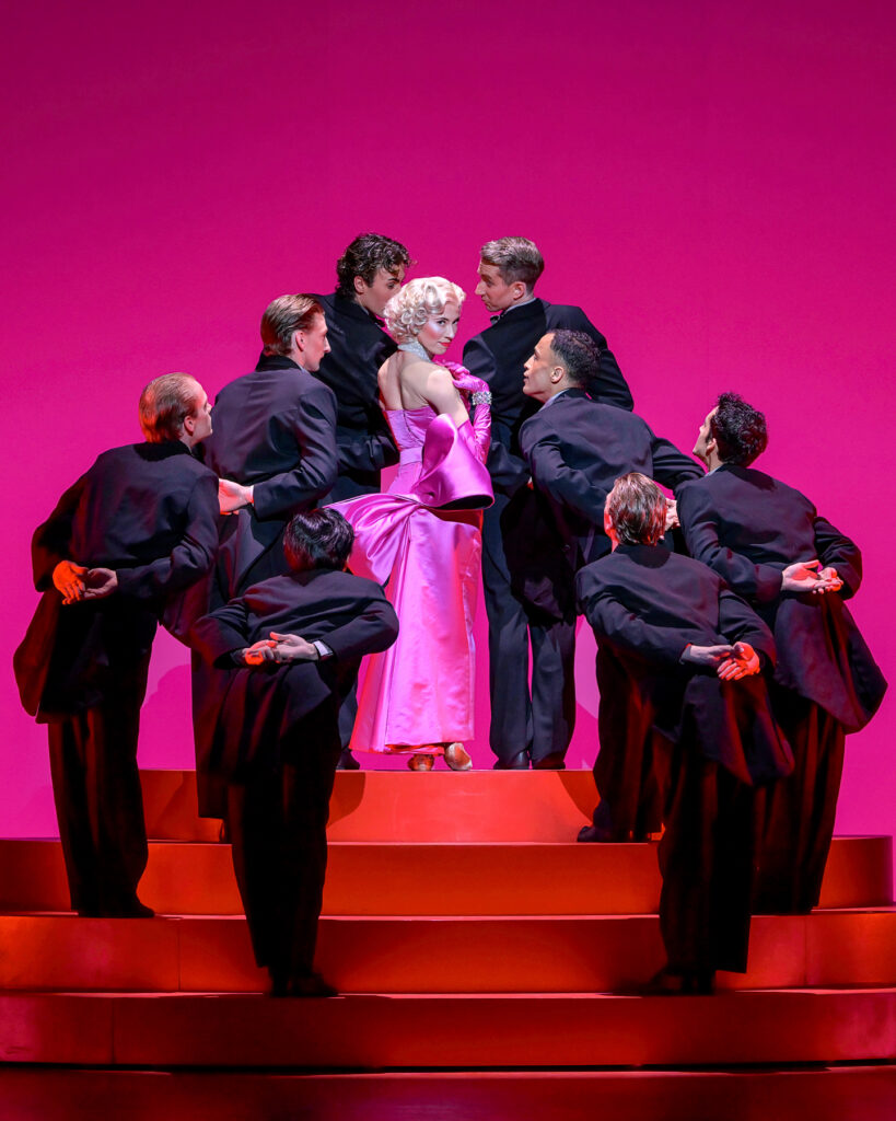 Charlotte Nash, costumed as Marilyn Monroe in a pink gown and elbow-length gloves and blonde wig, stands on top of a red staircase, surrounded by men in black tuxedos. She turns over her right shoulder and looks towards the audience seductively, touching her collarbone.