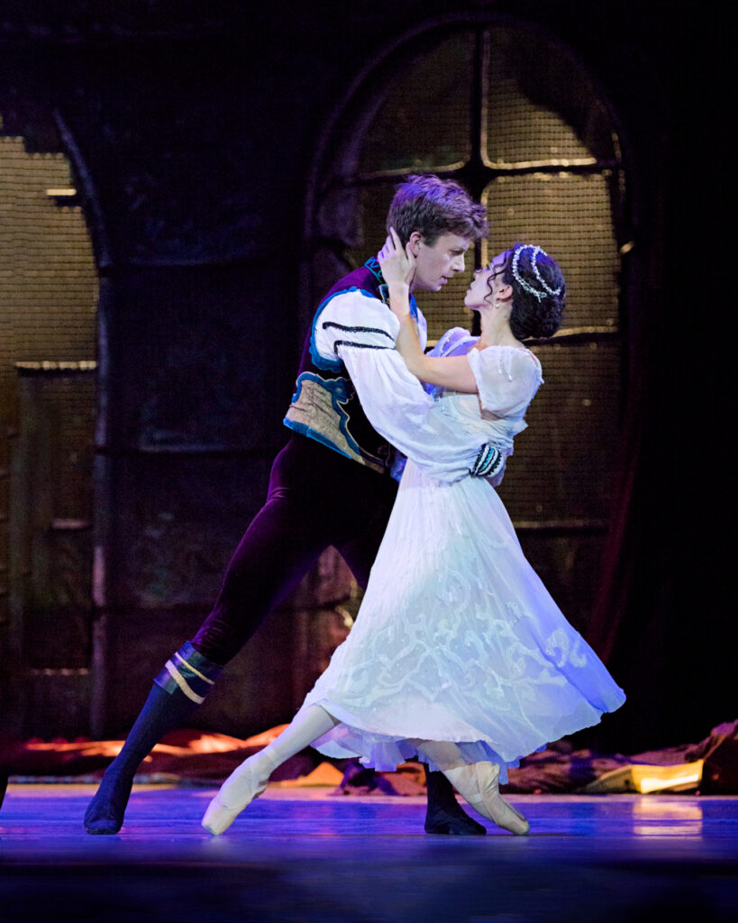 Charlotte Nash and John-Paul Simoens perform a pas de deux onstage. Simoens holds Nash by the waist as she touches his neck, her legs crossed with her left leg extended and her right leg in plié and on pointe. She wears a wedding dress and pink tights, while Simoens whites a white blouse with a dark burgundy vest and tights, and black boots.