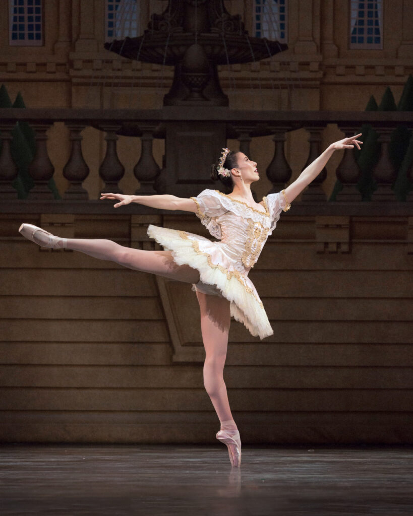 Charlotte Nash, wearing a tiara, white tutu with gold trim, and pink tights, performs a first arabesque on pointe on her left leg.