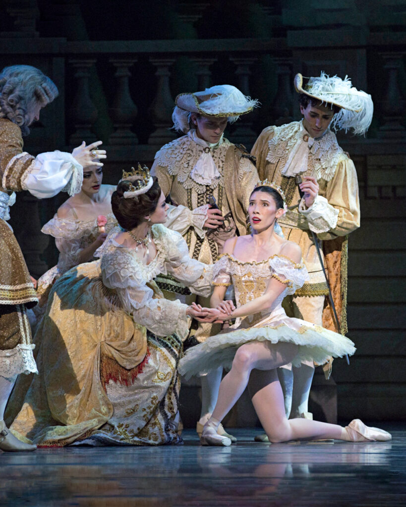 A ballerina wearing a white tutu with gold trim, is shown kneeling onstage, holding her left hand and looking up with a surprised expression. A dancer costumed as a queen kneels down next to her and looks at her with concern. A male dancer, costumed as noblemen, rushes towards her, while other dancers surround them,.