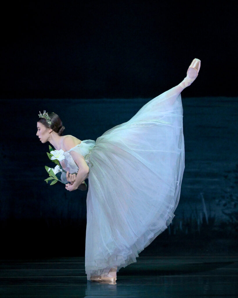 Charlotte Nash, wearing a long white Romantic tutu, flowered headpiece, pink tights and pointe shoes, does a penché arabesque on a darkened stage, holding two lilies close to her chest.