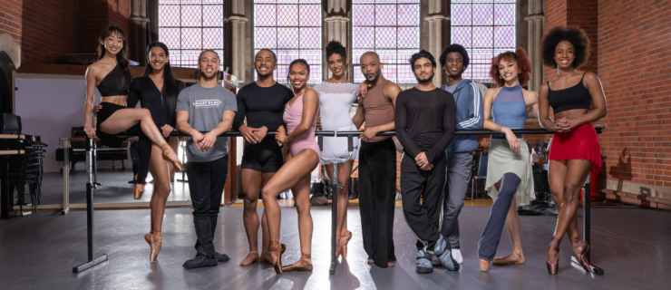 In a large ballet studio with four tall windows on the back wall, the company dancers of Ballet Black pose together around a ballet barre for a group photo.