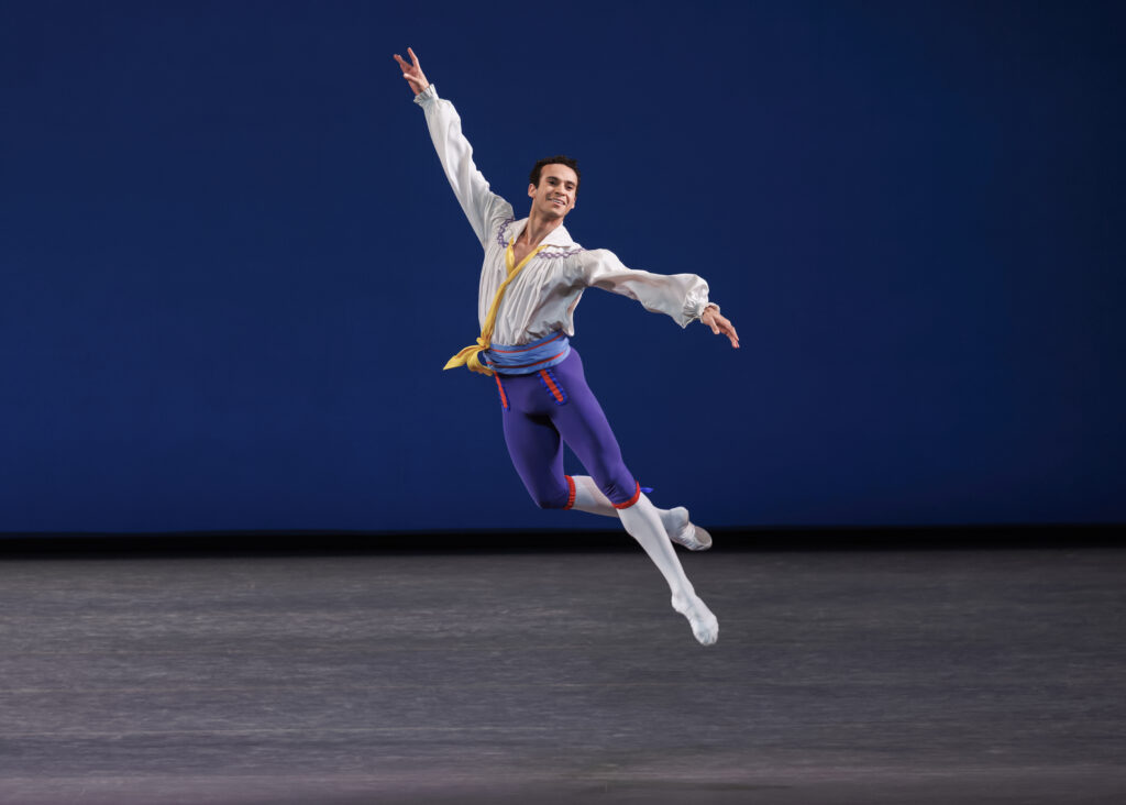 Onstage, a dancer jumps with one leg in a low attitude derriere, his arms extended in a high diagonal line. He smiles and wears a white tunic with puffy sleeves and a yellow sash, purple tights to the knee with white below, and a blue belt with red accents.