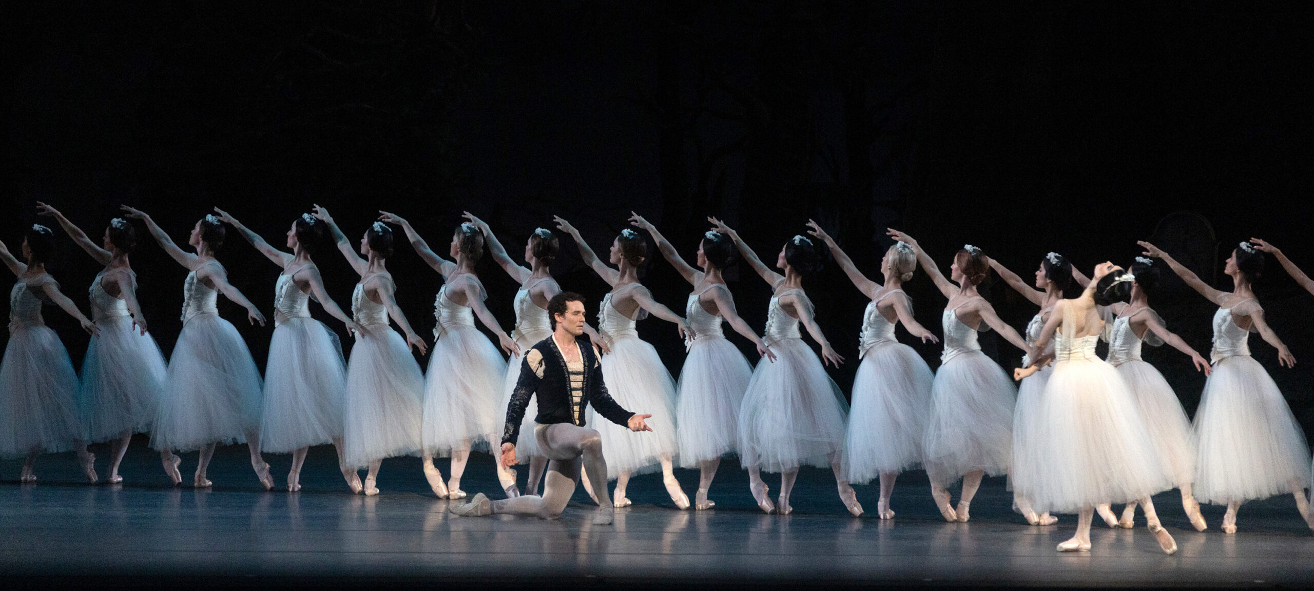 Onstage, a long line of corps de ballet dancers in white Romantic tutus extends from the top left corner to downstage right. In front of them, a dancer in a dark tunic and gray tights kneels, looking despondent, as another dancer stands facing him in tendu derriere, arching back with her arms crossed in the ballet pantomime for "death."