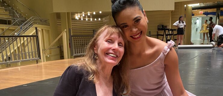 Patricia McBride and Angelica Generosa pose and smile, their heads leaning against each other. Genrosa wears a pink filmy dress, while McBride wears a short black jacket and white and black patterned dress.